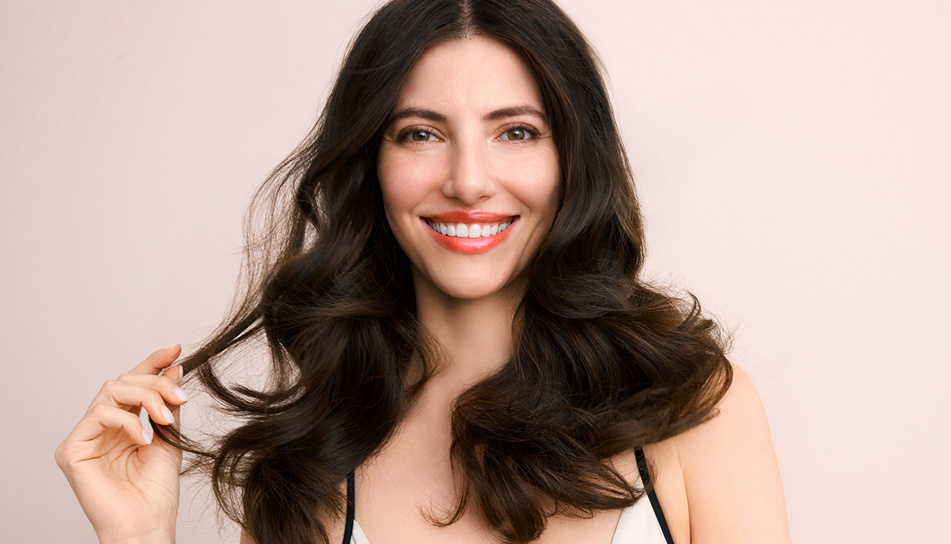 Smiling woman with long, dark, voluminous hair, gently holding a section of her hair against a light background
