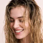 Smiling woman with wet, freshly washed hair against a soft light-pink background.