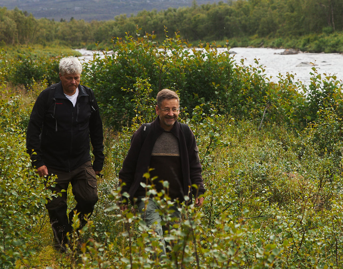 Two men walking in nature, surrounded by grass and flowers.