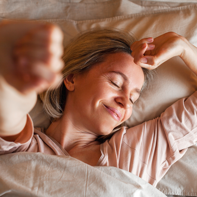 A woman waking up happily in bed, stretching and smiling as morning light fills the room.