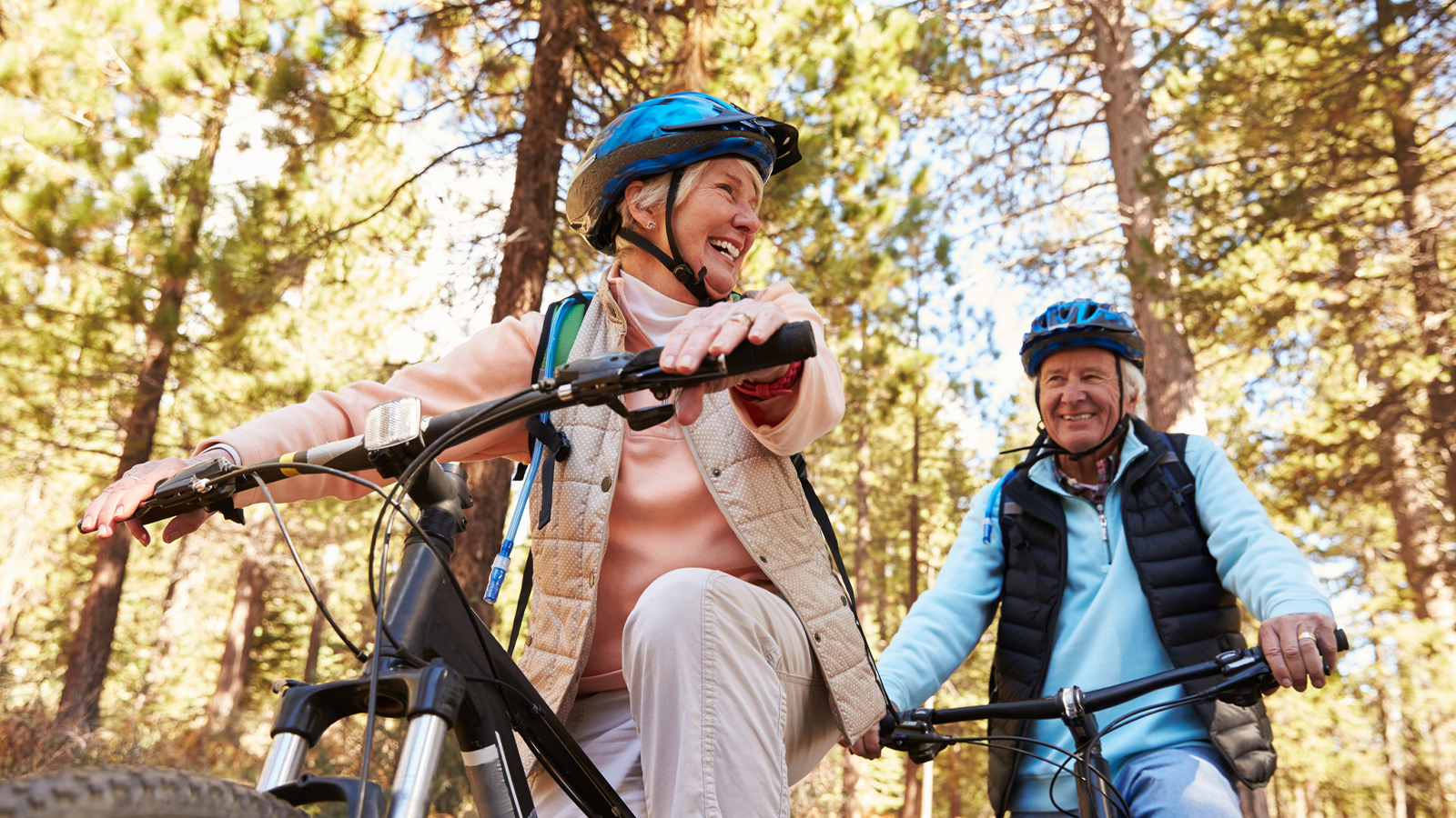 An older couple smiling as they ride bicycles together along a forest path surrounded by tall trees.