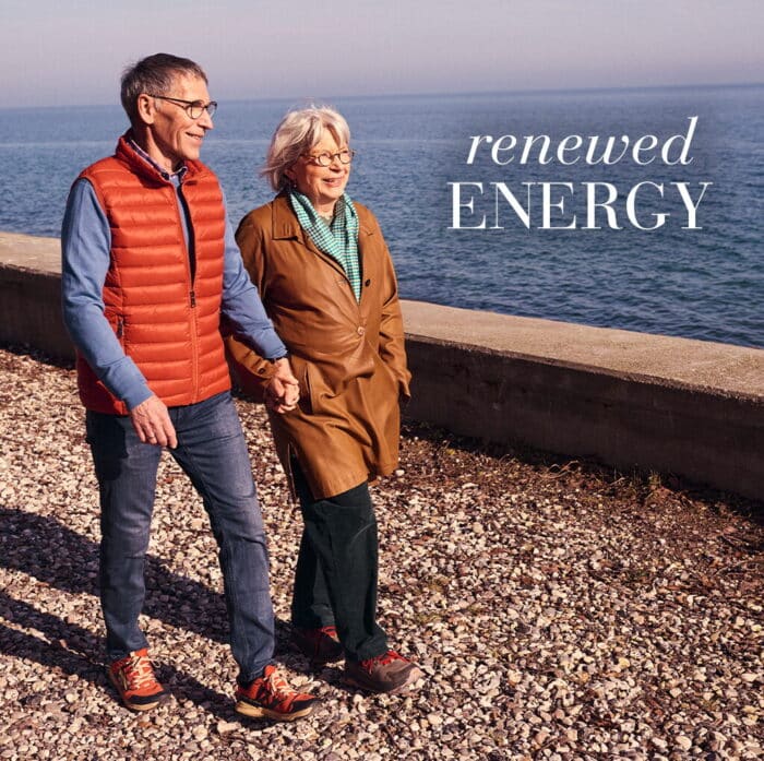 Older couple walking hand in hand along a rocky seaside path, with the text “renewed energy”.